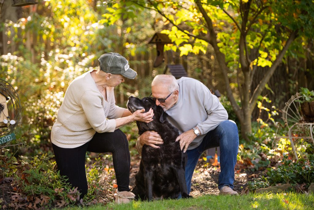 Elderly Man and Woman sitting petting dog in forest.